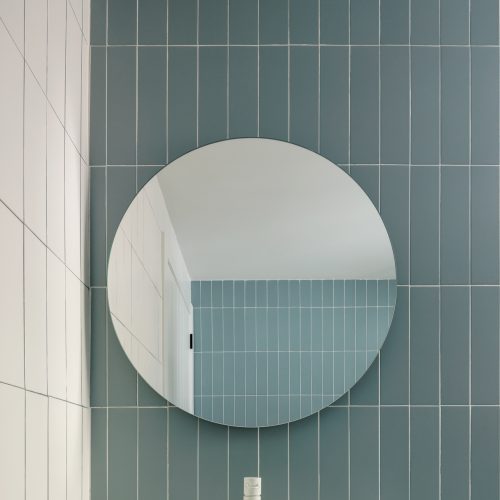 Arquitectura sostenible aplicada a baño con cerámica vertical azul, espejo circular y grifería blanca en vivienda reformada en Villa Muntanya