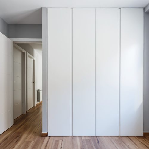 Dormitorio con armario empotrado blanco, radiador mural y ventana de madera, arquitecto interiorista en Valencia