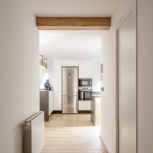 Vista desde el pasillo hacia cocina integrada con mobiliario blanco y encimeras oscuras proyectada por arquitecto obra nueva Valencia