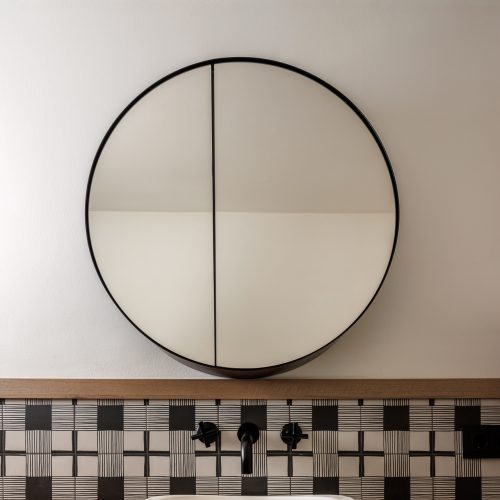 Arquitectura de interiores con lavabo sobre encimera de madera, espejo circular abatible y revestimiento cerámico gráfico en baño de Villa Muntanya