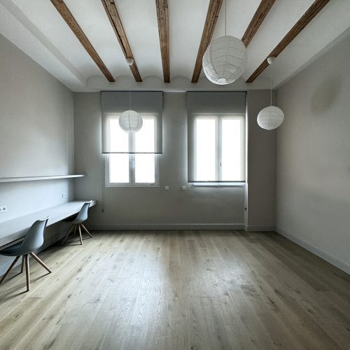 Espacio diáfano con vigas vistas, pavimento de madera y escritorio técnico diseñado por arquitecto residencial en vivienda reformada en Valencia