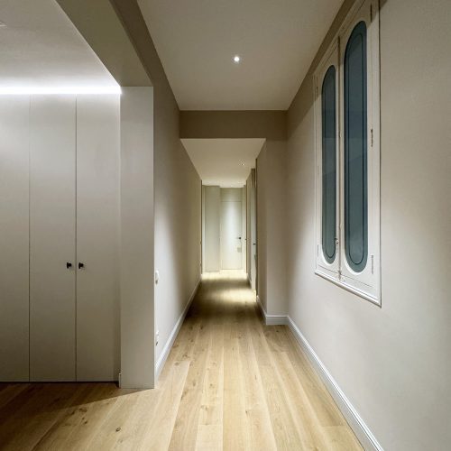 Pasillo con iluminación técnica, pavimento de madera y ventana restaurada como parte de reforma integral de vivienda en el Ensanche de Valencia