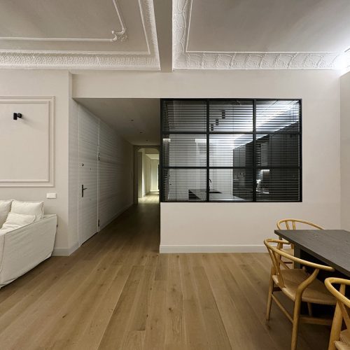 Zona de día con molduras restauradas, pavimento de madera y mampara metálica, ejemplo de arquitectura de calidad en vivienda reformada en Valencia