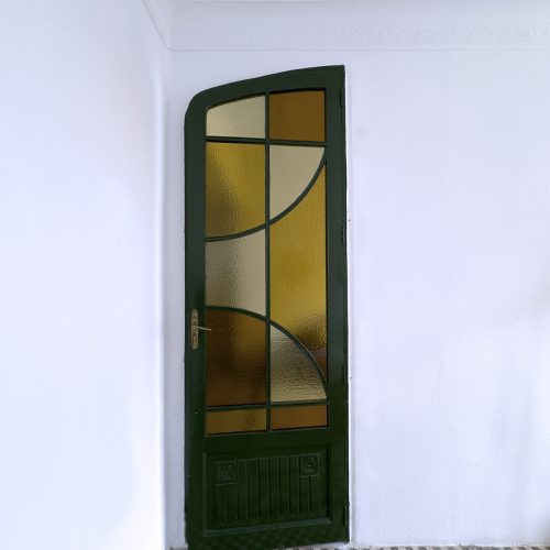 Puerta original restaurada con carpintería verde y vidriera ámbar sobre mosaico hidráulico en intervención de rehabilitación de viviendas en Ciutat Vella