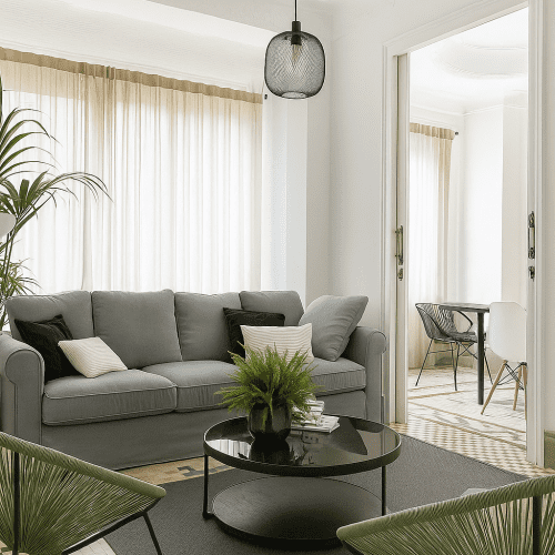 Salón luminoso con sofá gris verdoso, mesa circular de cristal y luz filtrada por cortinas, reformado por arquitectos Valencia.